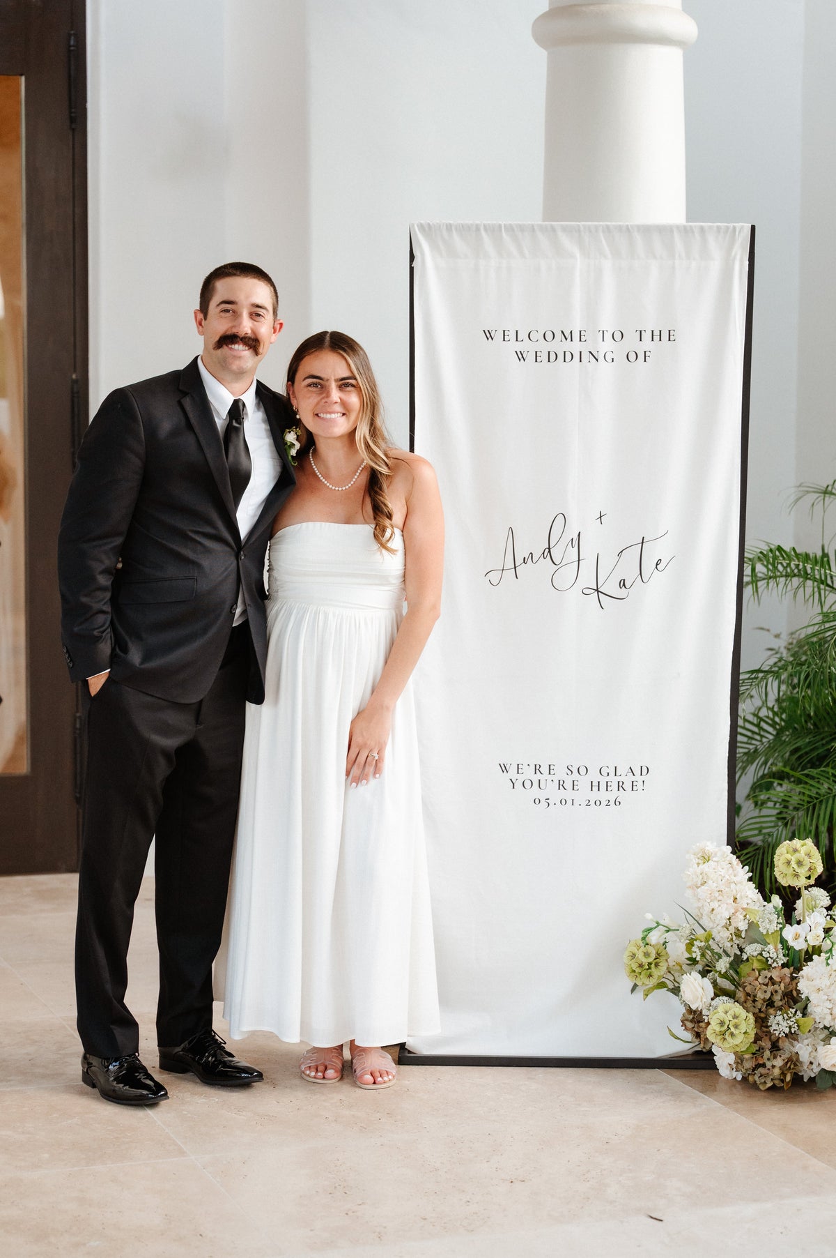 Bride and groom smiling beside a white fabric wedding welcome sign reading Andy + Kate at their Palm Springs wedding
