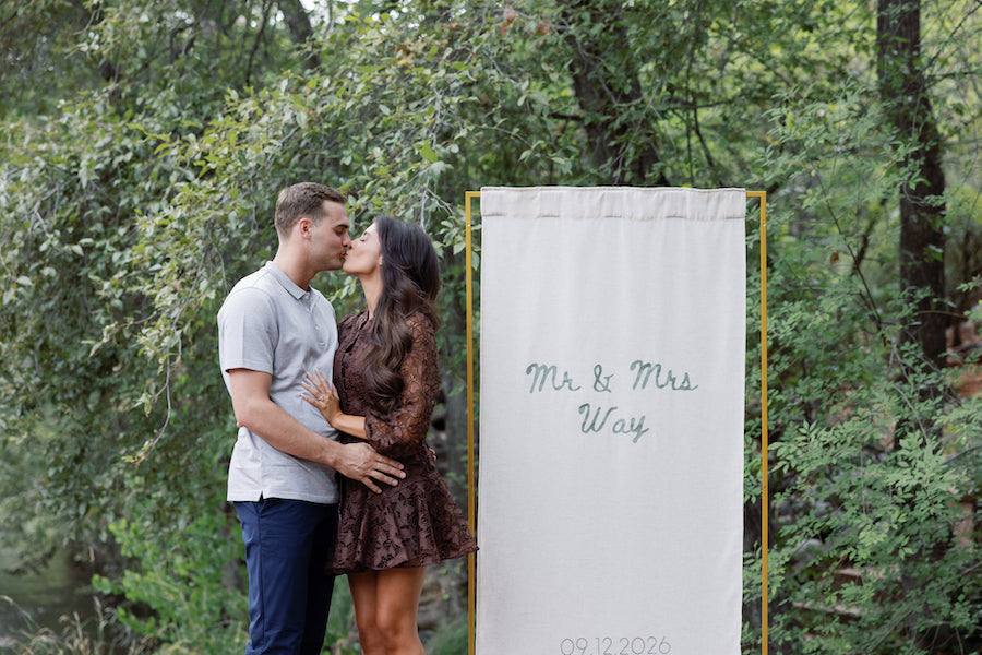 Couple kissing beside a custom fabric proposal sign reading Mr & Mrs on a gold stand among green trees in Sedona, Arizona