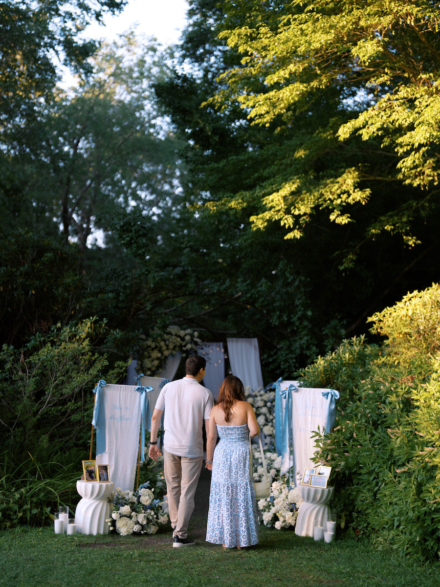 Couple walking hand in hand down a garden path lined with custom fabric proposal signs, blue satin ribbon and white florals in Long Island, NY