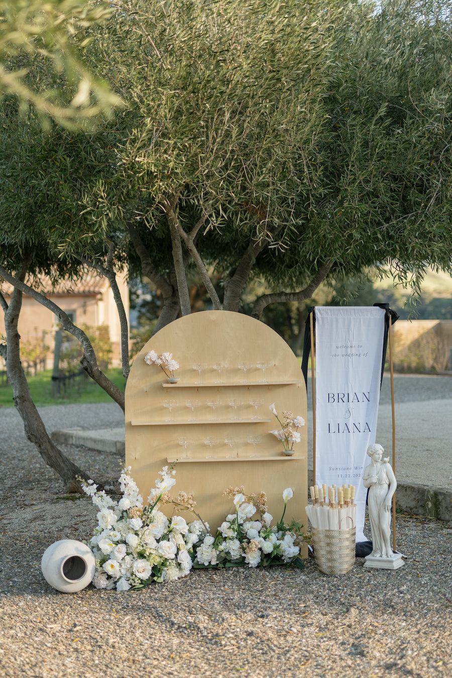 Custom fabric wedding welcome sign reading Brian & Liana on a gold stand beside an arched champagne display and white flowers under an olive tree at Sunstone Villa
