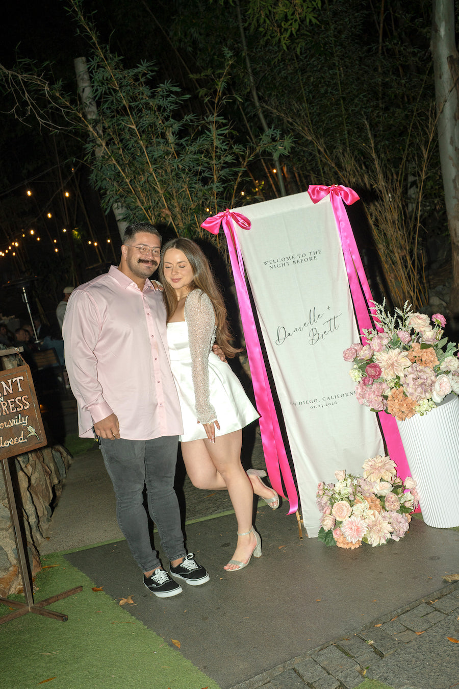Bride and groom posing beside a custom fabric wedding welcome sign with hot pink satin bows at their San Diego welcome dinner
