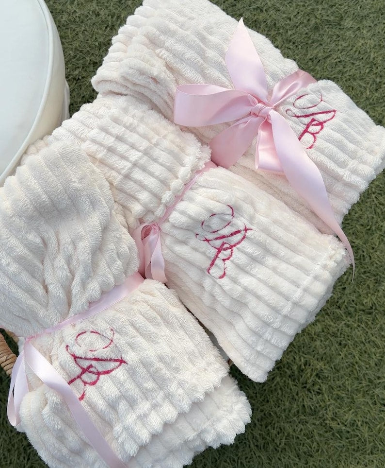 Three folded cream blankets tied with light pink satin ribbons, each with a dark pink embroidered cursive monogram