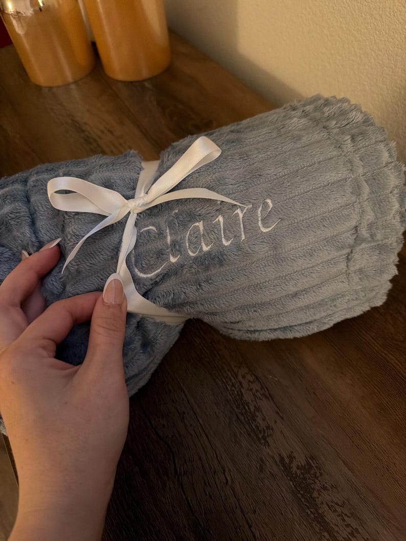 Rolled light blue plush blanket with a white embroidered name, tied with a white satin ribbon bow