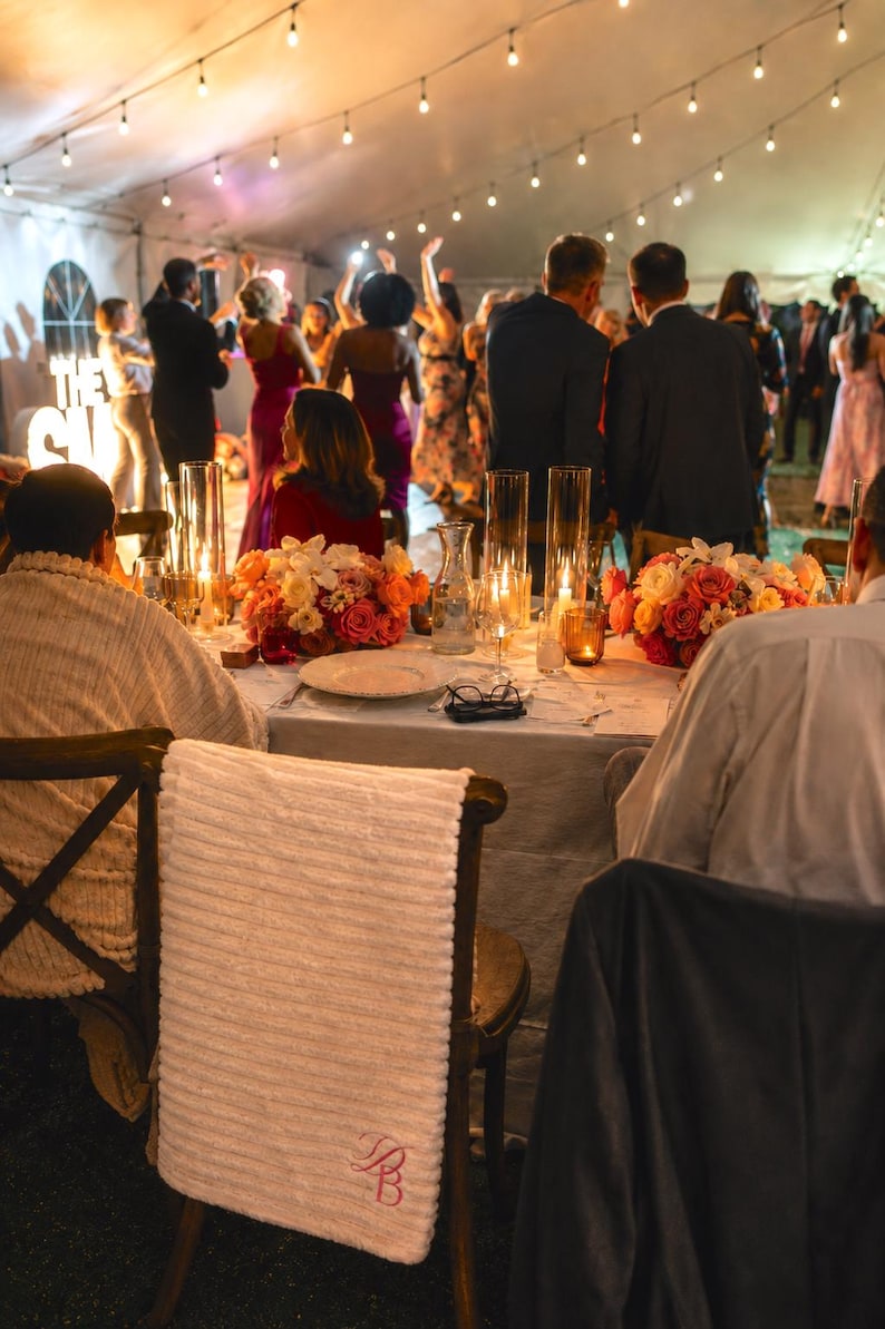 White textured blanket with a pink embroidered monogram draped over a wooden wedding chair beside floral centerpieces and string lights